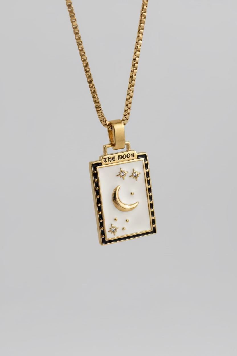 A gold-toned pendant necklace featuring a rectangular cream-colored face with a crescent moon and stars, displayed on a dark gray textured surface.
