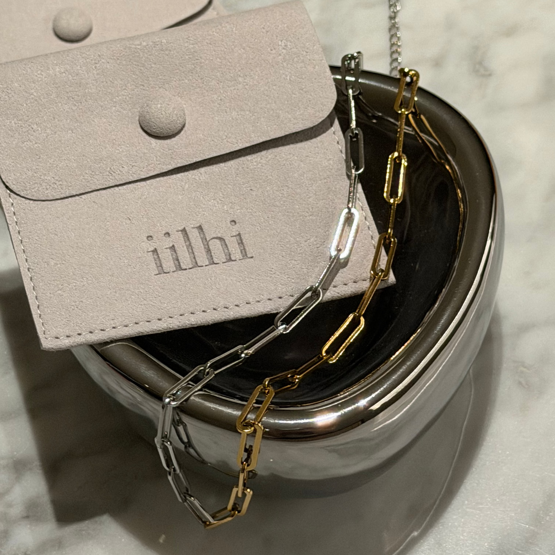 Jewelry box with gold and silver chain and a silver pouch on a marble surface.